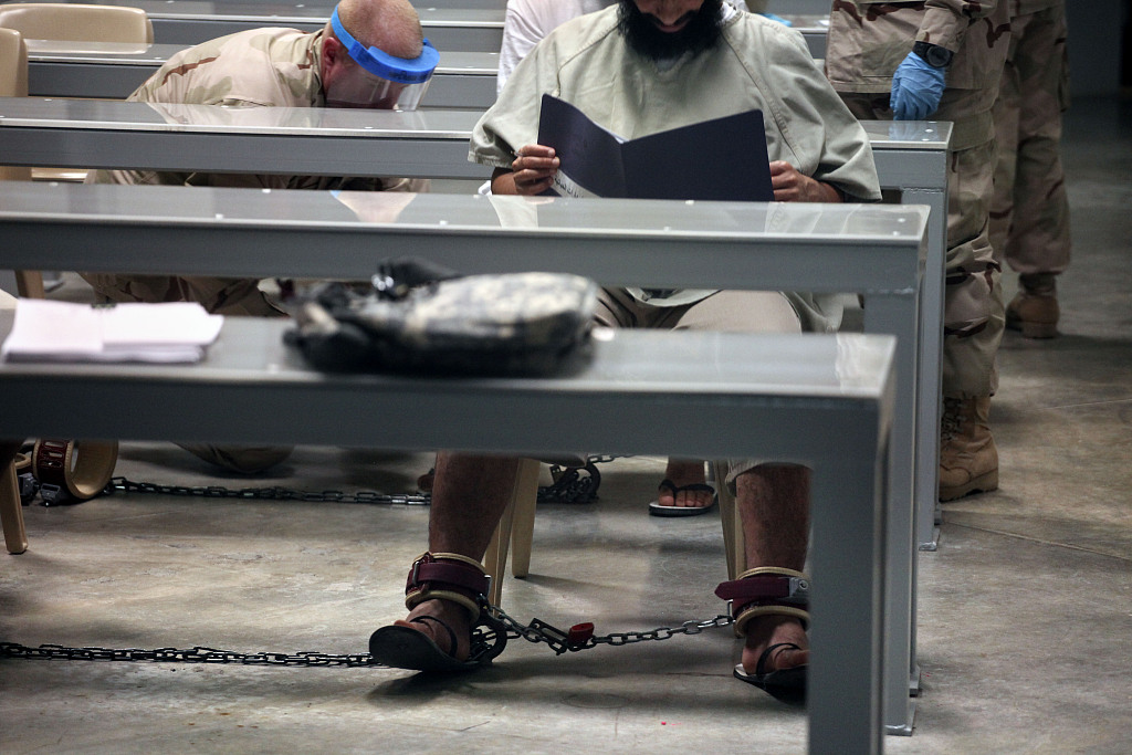 资料图：关塔那摩监狱内，一名囚犯正阅读材料，身后的看守将另一囚犯拷在地板上